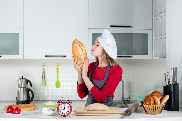 Front view pretty woman in cook hat and apron smelling bread in the kitchen