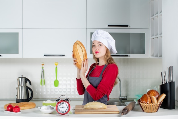 Вид спереди симпатичная молодая женщина в поварской шляпе и фартуке, держащая хлеб на кухне
