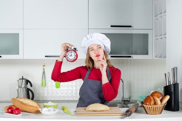 Front view pretty young woman holding up red alarm clock in the kitchen