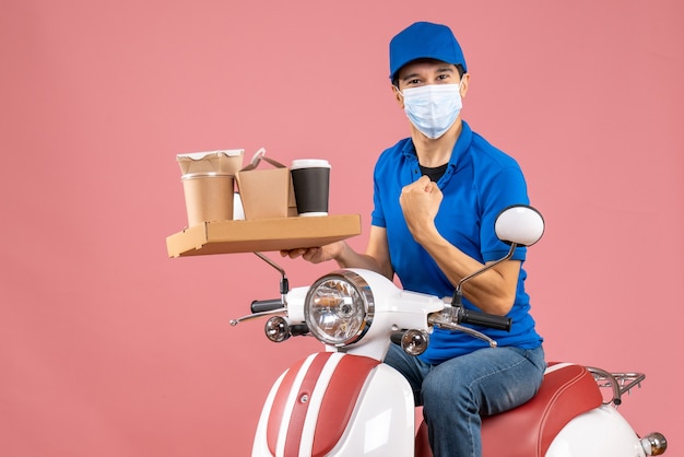 Front view of proud ambitious male delivery person in mask wearing hat sitting on scooter delivering orders on pastel peach background