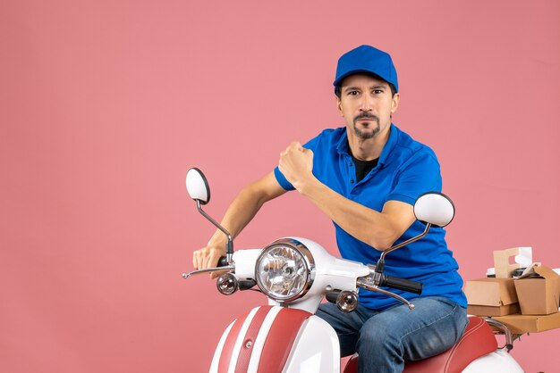 Front view of proud courier guy wearing hat sitting on scooter on pastel peach background