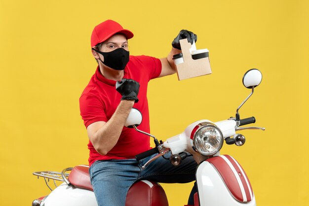 注文を示すスクーターに座っている医療マスクで赤いブラウスと帽子の手袋を着用している誇り高き宅配便の男の正面図