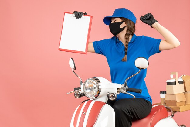 Front view of proud courier woman wearing medical mask and gloves sitting on scooter holding empty paper sheets delivering orders on pastel peach background