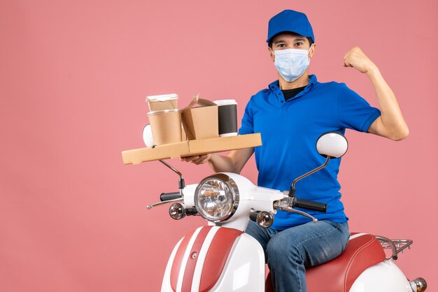 Front view of proud male delivery person in mask wearing hat sitting on scooter delivering orders showing his muscular on pastel peach background