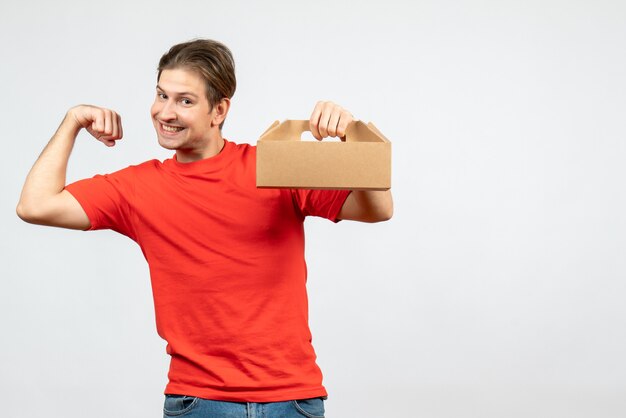 Front view of proud young man showing his muscular in red blouse holding box on white background