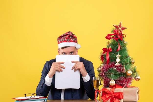 Вид спереди любопытного человека, держащего документы, сидящего за столом возле рождественской елки и подарков на желтом