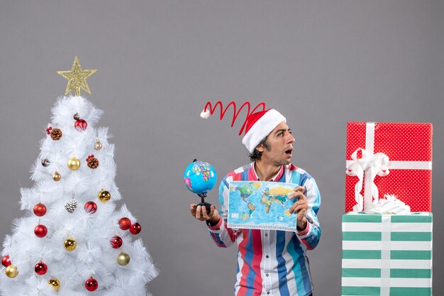 Вид спереди любопытный человек со спиральной весенней шляпой санта-клауса смотрит на подарки, держа карту мира и глобус