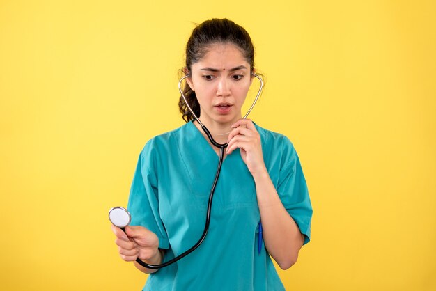 Front view prying woman doctor in uniform using stethoscope on yellow background