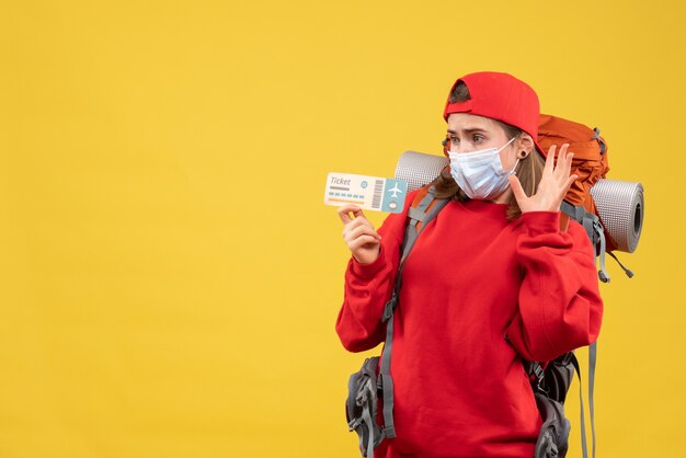 Front view puzzled female traveler with backpack and mask holding air ticket