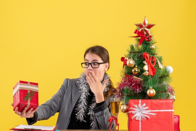 彼女のギフトのクリスマスツリーとギフトカクテルを見てテーブルに座っている眼鏡と正面図困惑した女の子