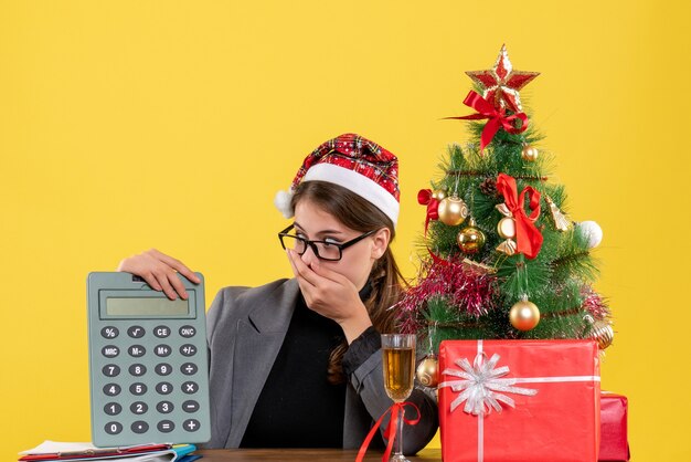 Front view puzzled girl with xmas hat sitting at the table loooking at calculator xmas tree and gifts cocktail