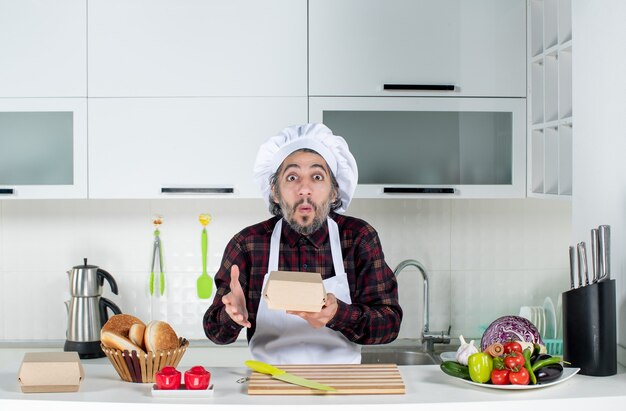 Front view of puzzled male chef in uniform holding box in the kitchen