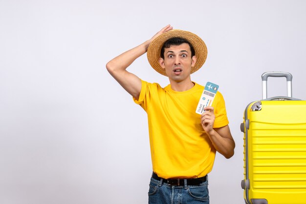 Front view puzzled young man in yellow t-shirt standing near suitcase holding up ticket