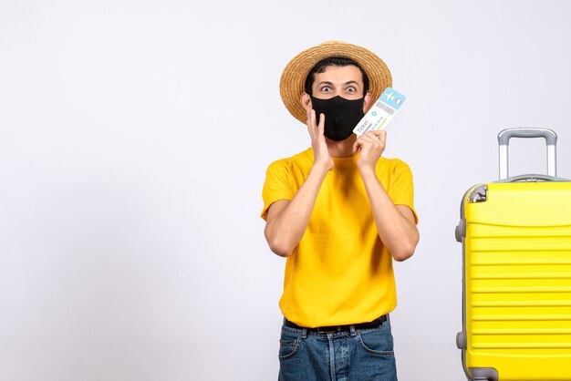 Вид спереди озадаченный молодой человек в желтой футболке, стоящий возле желтого чемодана с проездным билетом