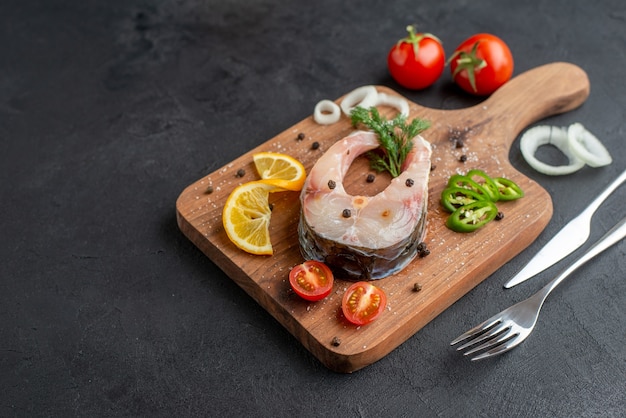 Front view of raw fish and fresh chopped vegetables lemon slices spices on a wooden board on the left side on black distressed surface