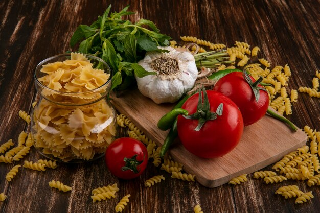 Front view of raw pasta in a jar with tomatoes garlic and chili peppers on a cutting board and with a bunch of mint on a wooden surface