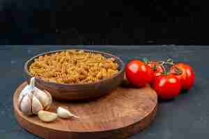 Foto gratuita vista frontale di pasta cruda in una ciotola marrone garlics su punte di legno e pomodori con steli su sfondo nero