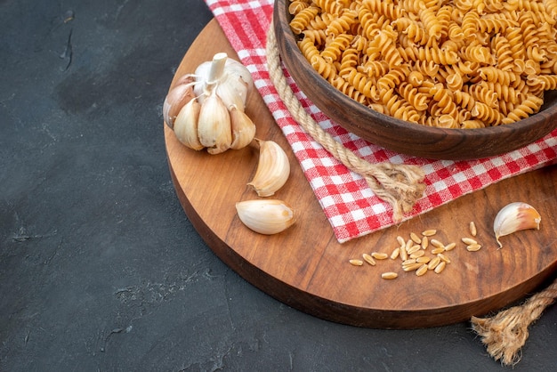 Front view of raw pastas in a brown bowl on red stripped towel garlics rice on round wooden board rope