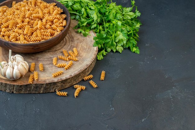 Front view of raw pastas inside and outside brown pot garlics green bundle on dark blue color background