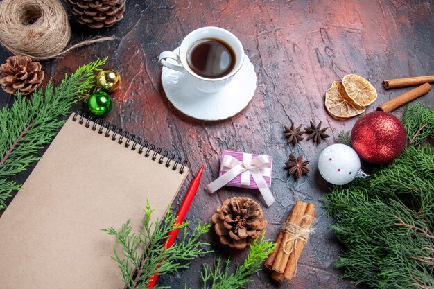 Front view red pen a notebook pine tree branches xmas tree ball toys and gift cinnamon anises cup of tea straw thread on dark red background