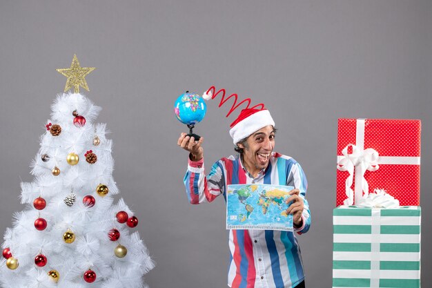 Front view rejoiced man holding world map and globe near xmas tree and presents