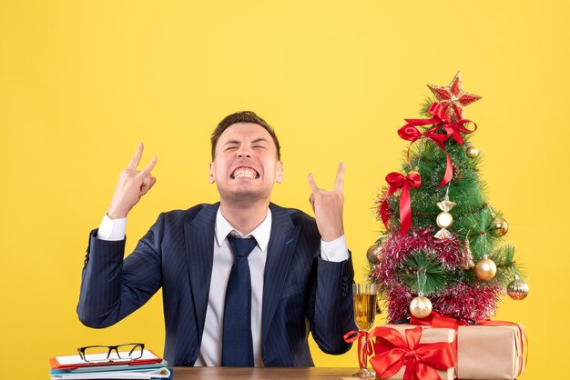 Vista frontale dell'uomo gioito che si siede al tavolo vicino all'albero di natale e regali su giallo