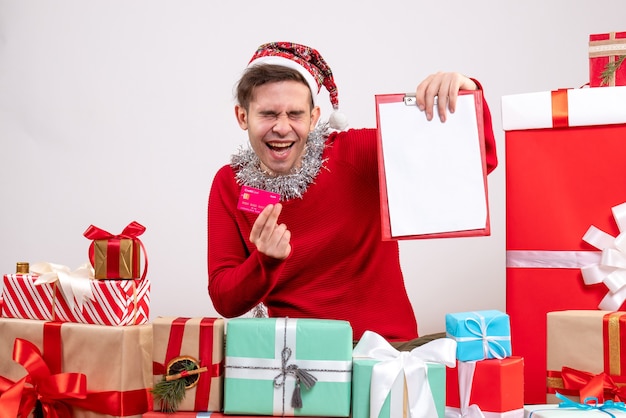 Front view rejoiced young man holding card and clipboard sitting around xmas gifts