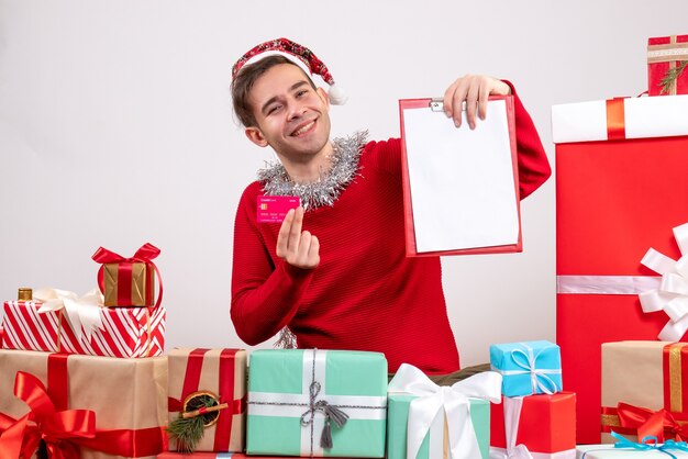 Front view rejoiced young man holding clipboard and card sitting around xmas gifts