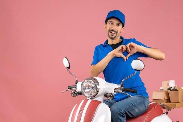 Front view of romantic delivery guy wearing hat sitting on scooter making heart gesture on pastel peach background