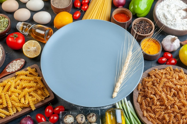 Front view round blue plate with raw pasta flour vegetables seasonings ands on dark 