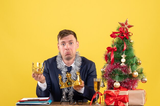 Front view of sad business man holding crowns sitting at the table near xmas tree and presents on yellow