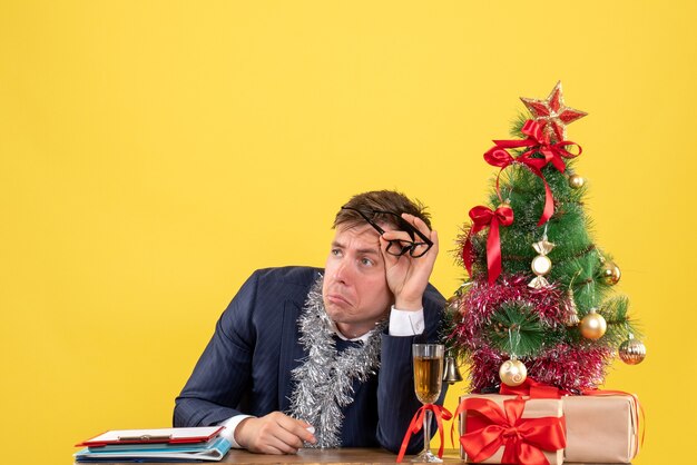 Front view of sad business man sitting at the table near xmas tree and presents on yellow