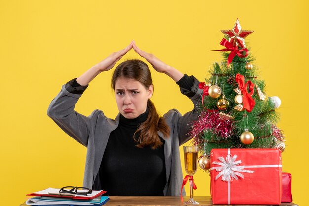 Front view sad girl sitting at the desk holding hands over head like a roof house near xmas tree and gifts cocktail