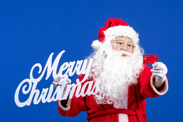 銀行カードと青い色の休日のプレゼントにメリークリスマスの書き込みを保持している正面のサンタクロース