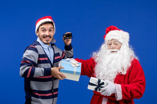 青い休日のクリスマスにプレゼントと銀行カードを保持している若い男性を保持している正面のサンタクロース