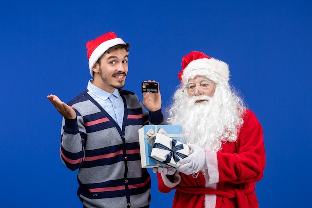 Front view santa claus holding presents and young male holding bank card on blue xmas holiday