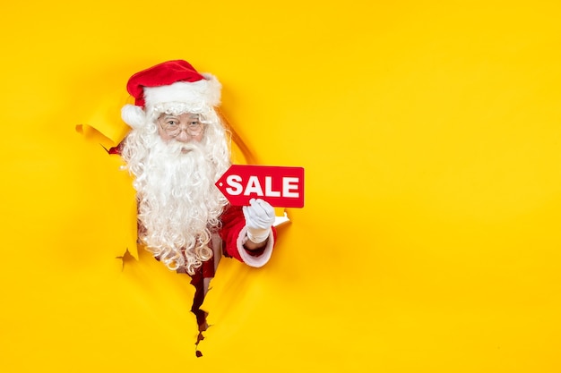 Free photo front view of santa claus looking through ripped paper yellow wall
