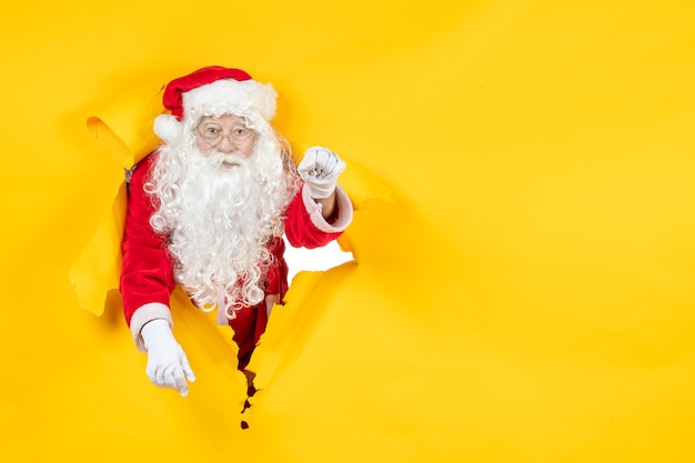 Free photo front view of santa claus looking through ripped paper yellow wall