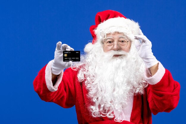 Front view santa claus in red suit holding black bank card on blue color holiday present xmas