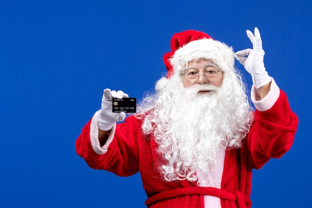 Front view santa claus in red suit holding black bank card on blue desk holiday present xmas color