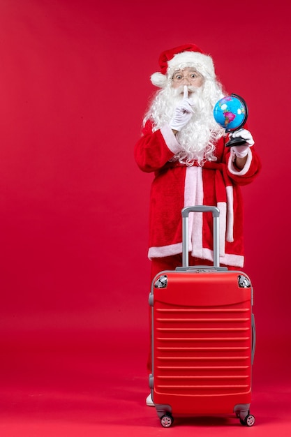 Front view of santa claus with bag holding little globe and preparing for trip on red wall