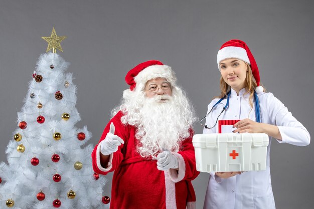 Front view of santa claus with female doctor who's holding first aid kit on a grey wall