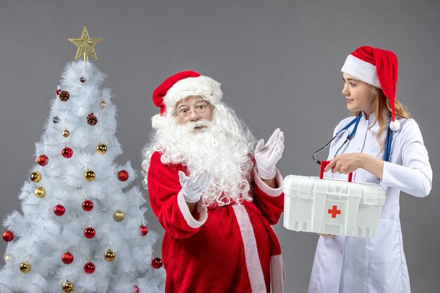 Front view of santa claus with female doctor who's holding first aid kit on grey wall