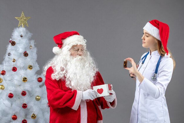 Front view of santa claus with female doctor who's holding little flask with pills on grey wall