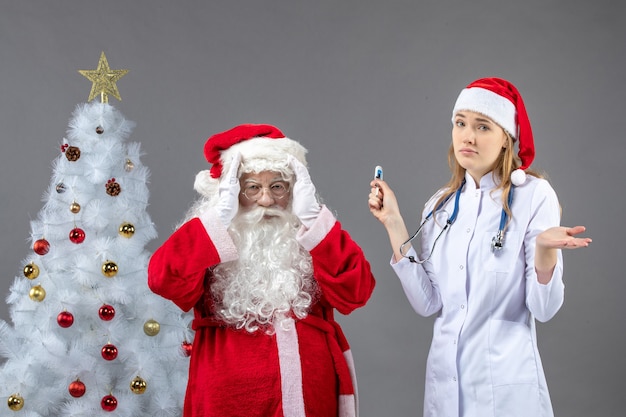 Front view of santa claus with female doctor who's holding temperature measuring device on a grey wall