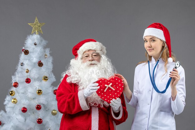 Front view of santa claus with female doctor who's observing present with stethoscope on grey wall