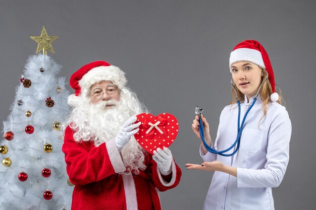 Front view of santa claus with female doctor who's observing with stethoscope on grey wall