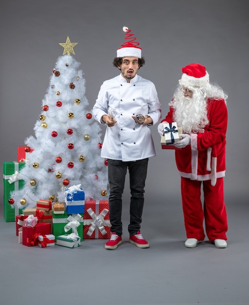 Front view of santa claus with male cook around christmas presents on a grey wall