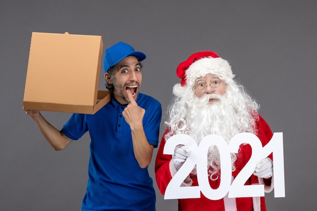 Front view of santa claus with male courier holding 2021 banner and food box on grey wall