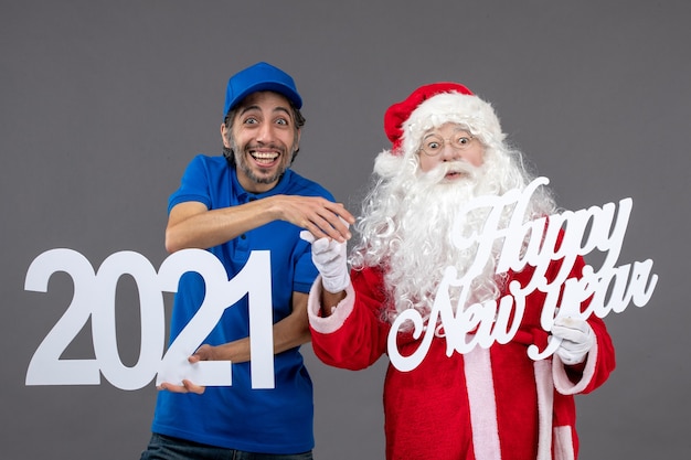 Free photo front view of santa claus with male courier holding and happy new year and 2021 boards on grey wall